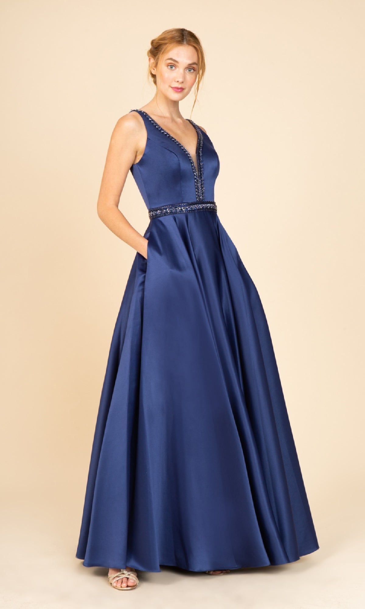 A woman models the Beaded-Waist Long Satin Prom Dress: Eureka 9010 in navy, featuring a sleeveless A-line silhouette with a deep V-neckline and beaded waist, standing against a plain beige background.